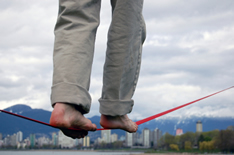 photo shows close of up a man's feet on a tightrope high above office buildings